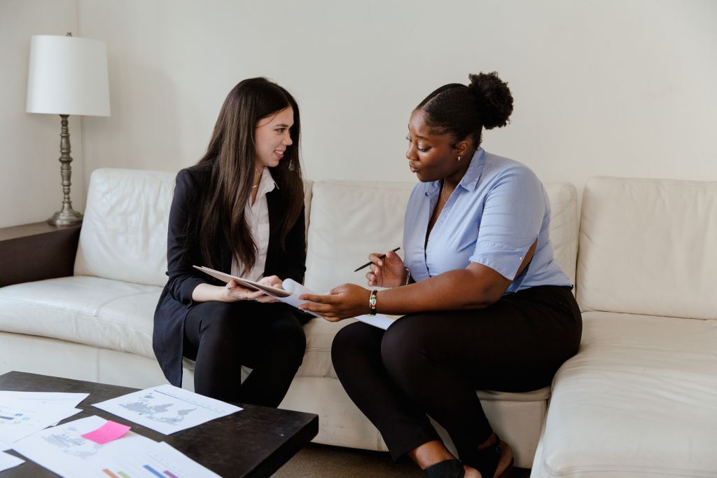 a black woman and a white woman in formal workwear during a SEO consultation, sat on a white sofa in an office